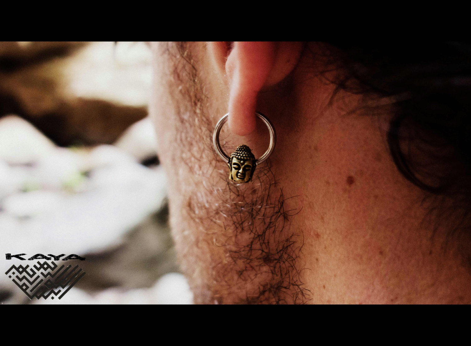 Close-up of a person's ear adorned with the BUDDHA Silver Ring with Gold Pendant by Kaya Movement. This versatile body jewelry, available in 18, 16, or 14 gauge, features a delicate Buddha pendant. The background is blurred with indistinct outdoor elements, and in the lower left corner the word "Kaya" and a geometric logo are visible.